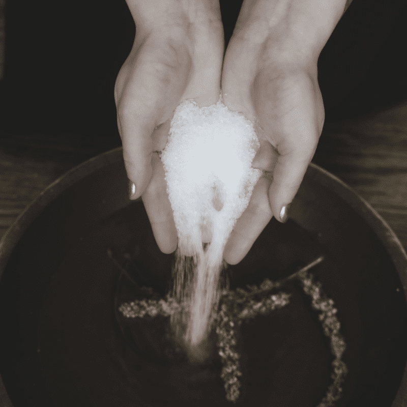 Hands pouring magnesium sulfate into a bowl with some herbs.