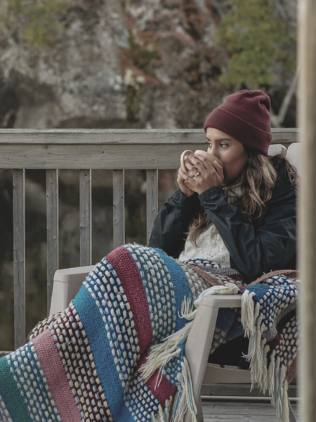 Woman sitting on a deck drinking tea with a blanket over her lap at sunset